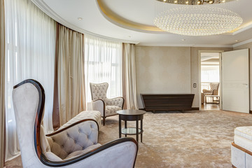 View of living room with two armchairs and rounded wooden table between. Interior of lighting room in beige and white colors. Big windows and luxury curtains.