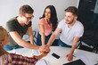 © estradaanton - For cheerful and happy coworkers stand around white table and look at each other. They hold hands together. People have materials on table.