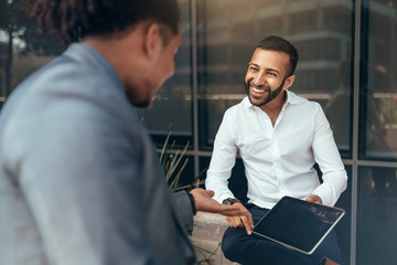 Two trendy businessmen talking and laughing casually outside and pointing to a digital tablet