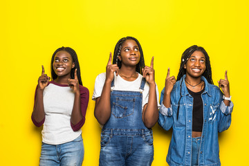 Wall Mural - Three beautiful smiling african women pointed up with hands looking up isolated over yellow background