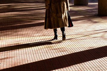 Wall Mural - Blurry silhouette shadow of legs of a woman in fur coat and high heels boots walking on a city arcade walkway