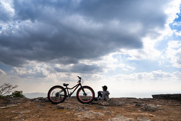 Wall Mural - A person sitting down beside a bicycle on rocky mountain looking out at scenic natural view and beautiful blue sky