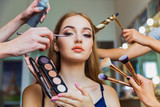 Portrait of the charming young woman who is sitting indoors and makeup artist doing makeup her and hairdresser doing the hairdo her