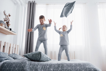 Happy kids playing in white bedroom. Little boy and girl, brother and sister play on the bed wearing pajamas. Nightwear and bedding for baby and toddler. Family at home