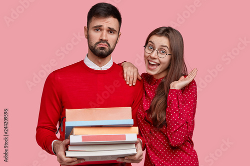 Fotografía Dissatisfied man with upset look, carries pile of books, tired of studying, happy Caucasian girlfriend expresses good emotions, stand closely, isolated over pink background