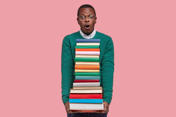 Emotional surprised black man looks with terrified expression, carries pile of textbooks, afraids of having many tasks to prepare, models over pink background, says omg, attends high school.