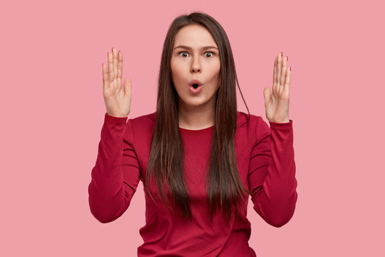 Photo of astonished brunette lady shows something big or huge with both hands, has surprised facial expression, wears red outfit, gestures in amazement, poses over pink background. Size concept