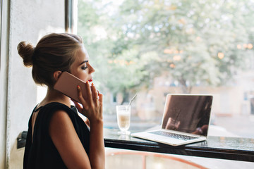 Portrait from side pretty woman in black dress in cafeteria. She is speaking on phone, looking on laptop