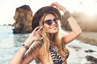 Portrait of attractive blonde girl with long hair posing on rocky beach. She wears bikini, hat, sunglasses. She is smiling.