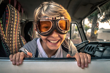 Blond smiling child looks out the window of vintage camper wearing aviator goggles sitting on colorful seat, puts his hands on the car door on the palms and sunset background. Vintage funny portrait