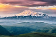 Beautiful view of Elbrus mount in the morning at sunrise with a dramatic cloudy sky and foggy green highland meadows. Summer mountain landscape. North Caucasus, Karachay-Cherkess Republic, Russia