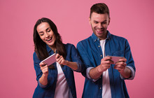 Young excited couple standing isolated over pink background in casual wear, playing games on mobile phones
