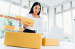 © Mymemo - Business owner concept. Young Asian casual woman smiling happy using laptop working checking customer's order and products packaging for delivery in modern home office