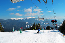 People In Colorful Sportwear Are Skiing Down The Slope Under Below Chairlift With Skiers And Snowboarders Lifting To Top Of Mountain In Ukraine. Landscape Of Winter Hills In Sunny Day. Sport Concept.