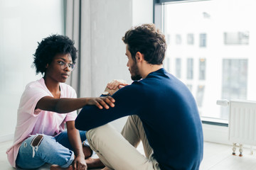 Young African woman and Caucasian man sitting near the window, staring at each other meditating together, free their minds from thoughts and worries.