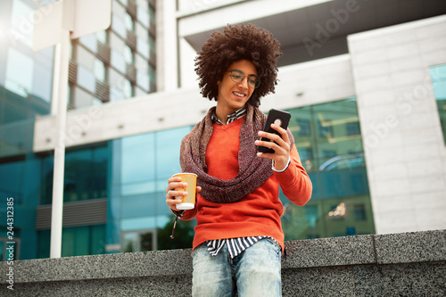 Foto A handsome curly-haired guy of mixed race is dressed in a trendy youth-style scarf and sweater writes a message in a teleone against the background of office buildings