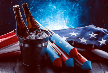 two bottles of beer in an ice bucket with the american flag lying nearby and rockets for fireworks. 