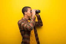 Young African American Man On Vibrant Yellow Background And Looking For Something In The Distance With Binoculars