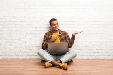 African american man sitting on the floor with his laptop holding copyspace imaginary on the palm to insert an ad