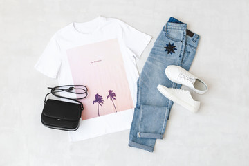 Wall Mural - White t-shirt with pink print, blue jeans, small black cross body bag and white sneakers on grey background. Overhead view of woman's casual outfits. Flat lay, top view. Trendy hipster look.