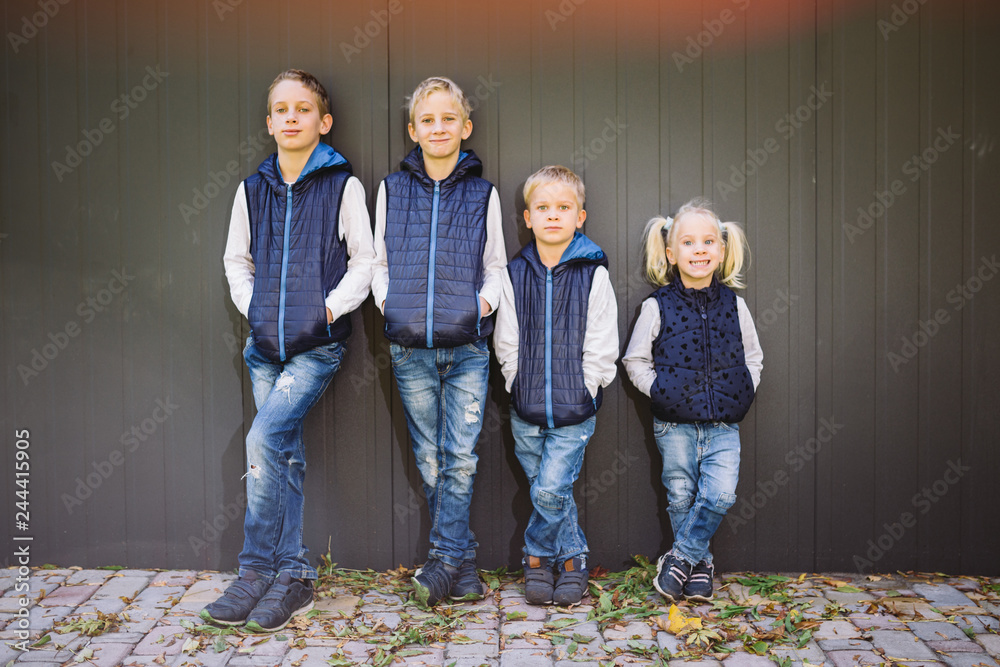 Funny Caucasian big family of three brothers and sister posing standing on growth background of wall in full growth. Equally stylishly dressed in blue vests and jeans. Theme girl in male circle - obrazy, fototapety, plakaty 
