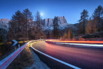 Wall Mural - Blurred car headlights on winding road at night in autumn. landscape with asphalt road, light trails, mountains, trees, and blue sky with moon at dusk. Roadway in Italy. View with highway and rocks