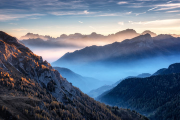 Mountains in fog at beautiful sunset in autumn in Dolomites, Italy. Landscape with alpine mountain valley, low clouds, trees on hills, village in fog, blue sky with clouds. Aerial view. Passo Giau