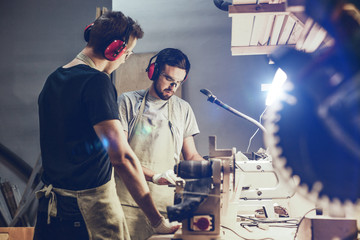 Wall Mural - Two attractive young men in aprons and earmuffs standing near workbench in brightly illuminated workshop and working with wood together