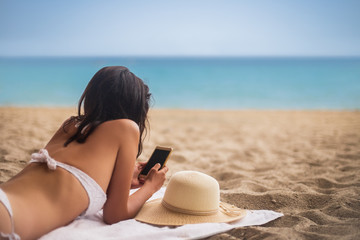 Wall Mural - Young beautiful woman lying on tropical beach and using mobile phone