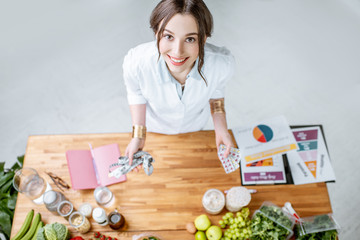 Wall Mural - Portrait of a young woman nutritionist in medical uniform standing near the table full of various healthy products indoors