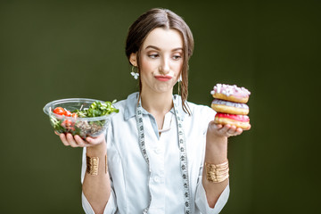 Wall Mural - Young woman nutritionist looking on the donuts with sad emotions choosing between salad and unhealthy dessert on the green background