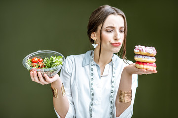 Young woman nutritionist looking on the donuts with sad emotions choosing between salad and unhealthy dessert on the green background