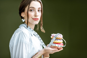 Wall Mural - Portrait of a sad woman nutritionist in medical gown with donuts and tape measure on the green background. Unhealthy eating and adiposity concept
