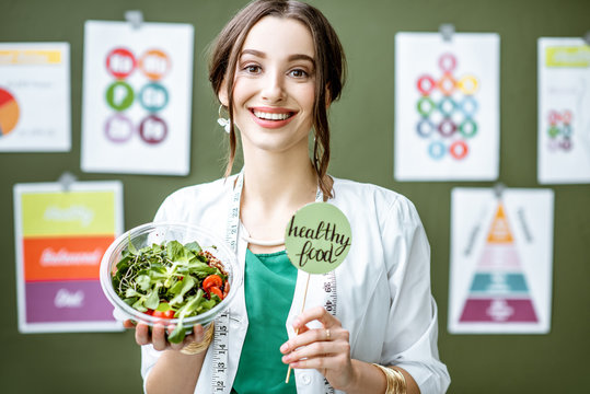 Portrait of a woman dietitian in medical gown standing with salad on the green wall background with drawings on a topic of healthy food indoors