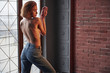 Fashion photo. Hot young blonde with bare chest and jeans stands against the window