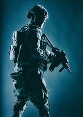 Army soldier in helmet, battle uniform with tactical ammunition, hiding face behind mask and glasses, looking back over shoulder, sneaking in darkness with service rifle in hand, low key, studio shoot