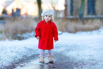 Outdoor winter portrait of little cute toddler girl in red coat and white fashion hat barret. Healthy happy baby child walking in the park on cold day with snow and snowfall. Stylish clothes for kids.