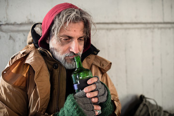 Wall Mural - A front view of homeless beggar man sitting outdoors, holding bottle of alcohol. Copy space.