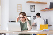 © Rithor - Fatigue from work. Tired young woman sitting at working table with glasses looking at the computer rubbing her aching head against the background of blurred man
