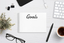 Creating Goals List On Notepad On Office Desk Surrounded With Office Supplies. White Wooden Work Desk.