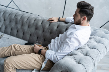 Wall Mural - Feeling lonely. Stylishly dressed young man holding a glass while sitting on sofa.