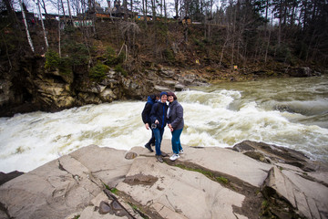 Family of three walk near river after nature hiking. People resting on rock in forest river enjoying autumn day. Active family life in nature.