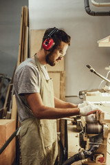 Wall Mural - Side view of handsome craftsman in earmuffs and goggles standing near workbench and using professional belt sander