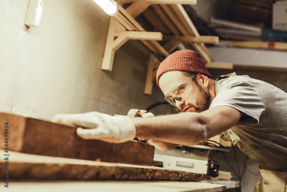 Attractive guy in protection goggles working with lumber plank on workbench in professional joinery - obrazy, fototapety, plakaty 