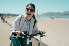 Girl Backpacker Rent Bike Travel Tour In San Francisco California Usa. Young Asian Woman Face Camera Smiling Cheerfully While Riding Bicycle On Beach. Golden Gate Bridge With Blue Sky In Background.