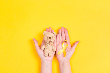 girl hands holding children's toy with a childhood cancer awareness yellow ribbon on yellow backgrou