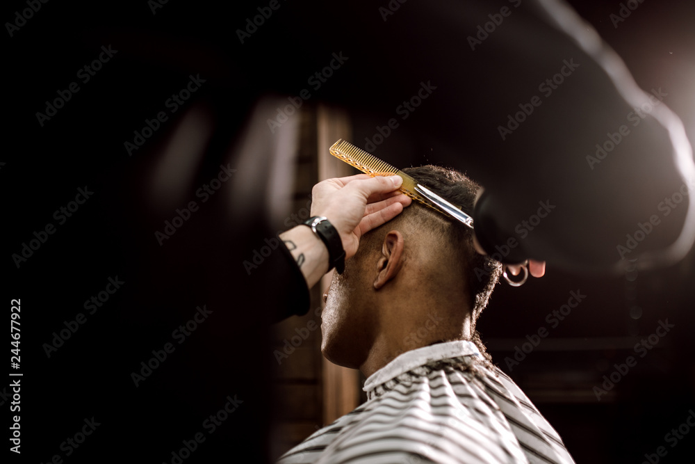 The barber scissors hair on the sides for a stylish black-haired man in the barbershop. Men's fashion and style - obrazy, fototapety, plakaty 