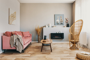 Wicker peacock chair, wooden coffee table and pink couch in grey living room interior