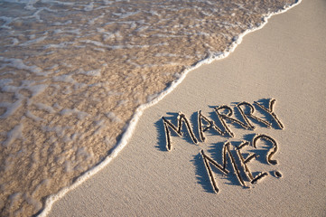 Marry Me? marriage proposal message handwritten in smooth sand with wave coming ashore on the beach