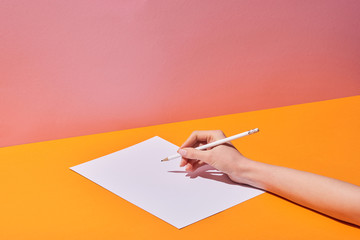 cropped view of woman writing on paper on yellow desk and pink background